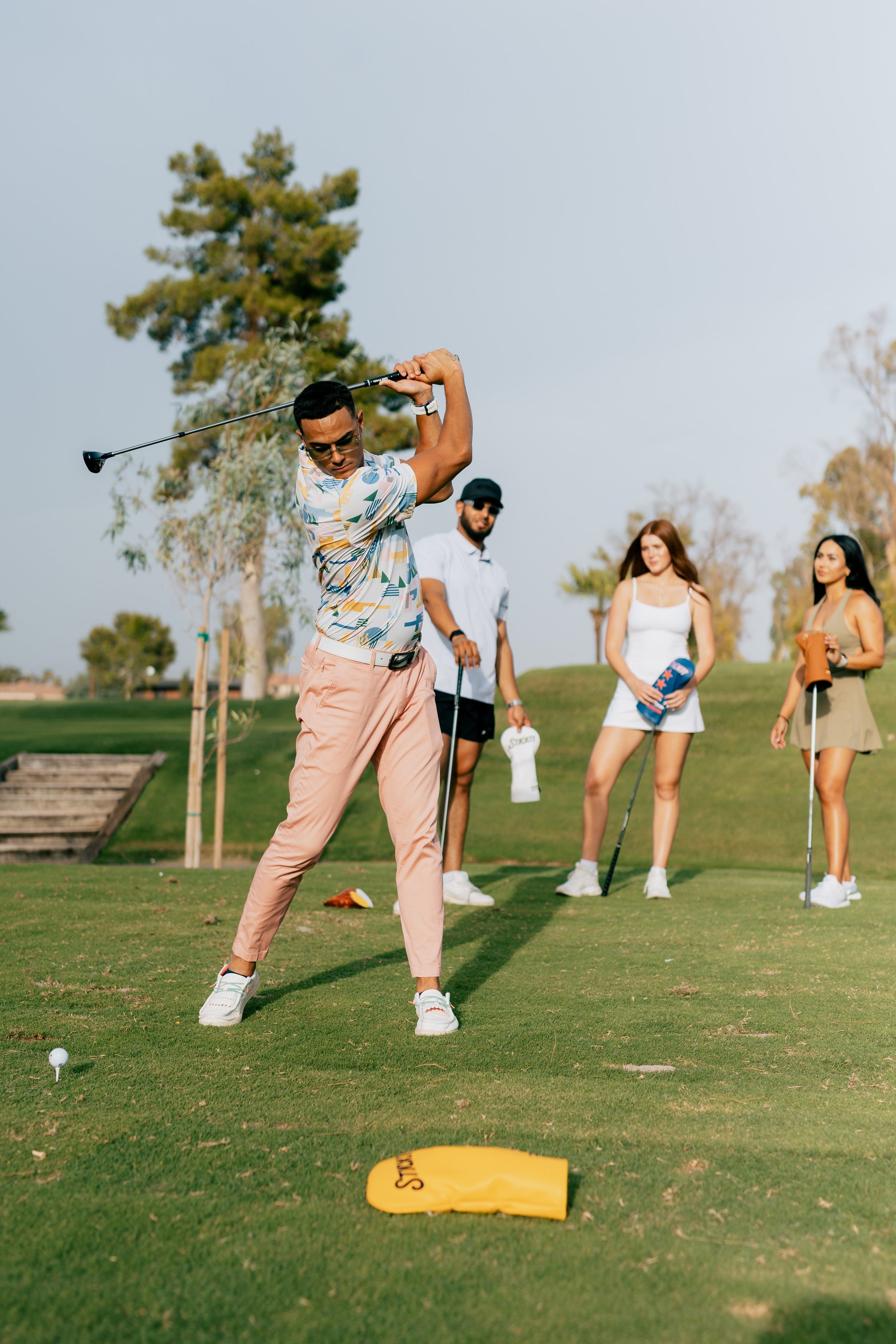 Group of golfers on the course with STICKIT gear—golfer swinging driver while friends watch with golf towels, headcovers, and accessories on a sunny day.