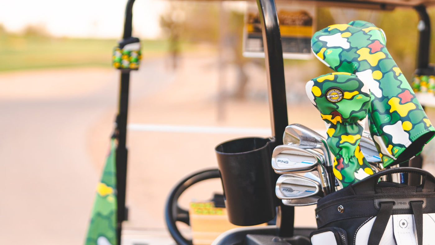 Green camo golf club covers with yellow accents, placed on a black golf bag in a cart on a sunny course.