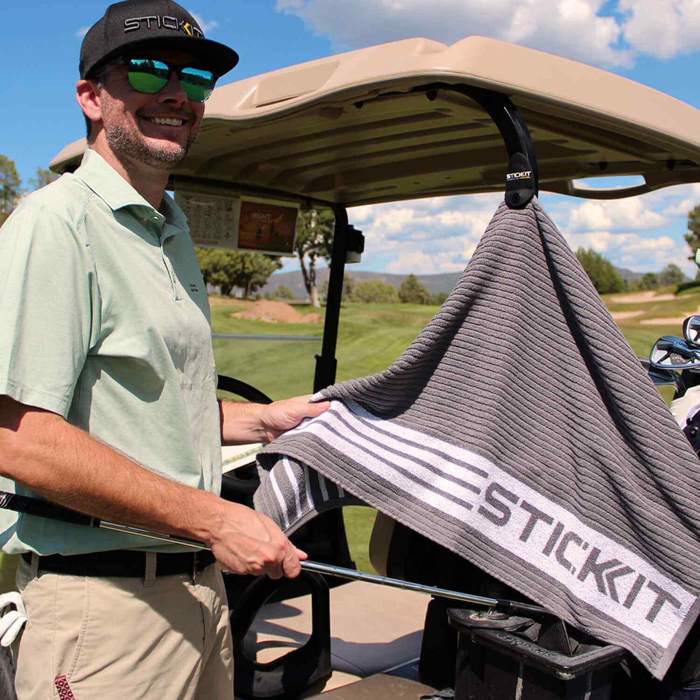 Gray golf towel with white stripes, branded "STICKIT," displayed by a golfer wearing a light green polo and black cap.